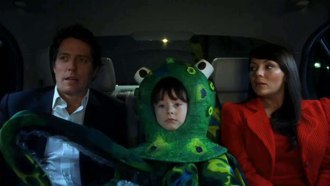 Boy in green octopus costume sits between Hugh Grant and Martine McCutcheon in Love Actually. 