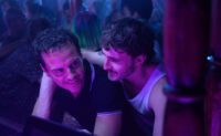 Adam and Andrew embrace, bathed in the purple light of a club.