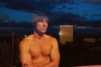 Adam, shirtless, looks out of his apartment window