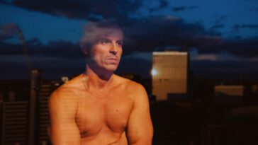 Adam, shirtless, looks out of his apartment window