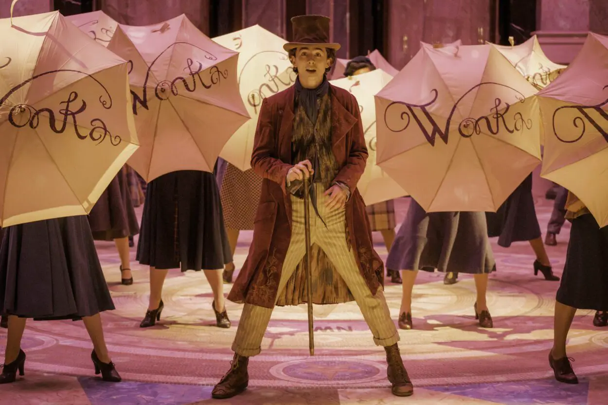 Willy Wonka (Timothée Chalamet) dances with a top hat and a cane while multiple dancers stagger him all with umbrellas open that have Wonka written on them.