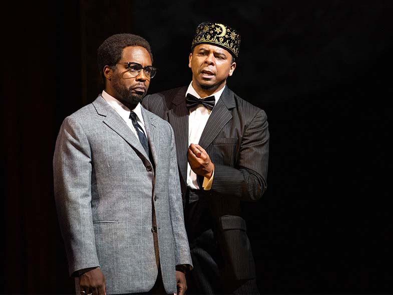 Malcolm X and a man in a crown stand on stage