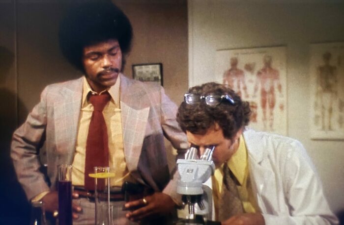 Richard Lawson and Gary Chason as Lt. Valentine and Lab Technician in Sugar Hill (1974). Screen capture off Amazon. An American International Picture/MGM. Afro sporting investigating detective Lt. Valentine waits for results from the Lab Technician inspecting evidence under a microscope.