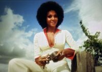 Marki Bey as Diana "Sugar" Hill and Langston in Sugar Hill (1974). Screen capture off Amazon. An American International Picture/MGM. Sugar Hill sits on a wooden fence wearing a white and red track suit, hair in an afro, playing with an old iron shackle.