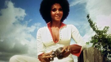 Marki Bey as Diana "Sugar" Hill and Langston in Sugar Hill (1974). Screen capture off Amazon. An American International Picture/MGM. Sugar Hill sits on a wooden fence wearing a white and red track suit, hair in an afro, playing with an old iron shackle.