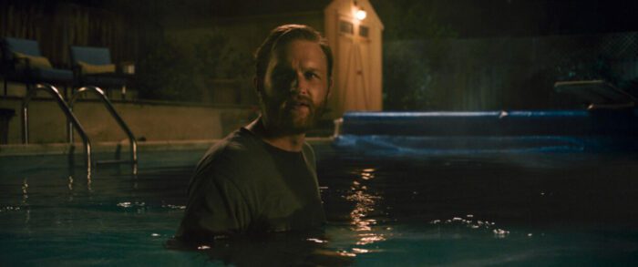 Wyatt Russell as Ray Waller in Night Swim, directed by Bryce McGuire. © 2023 Universal Studios. All Rights Reserved. Ray Waller walks around inside a dark pool fully clothed.