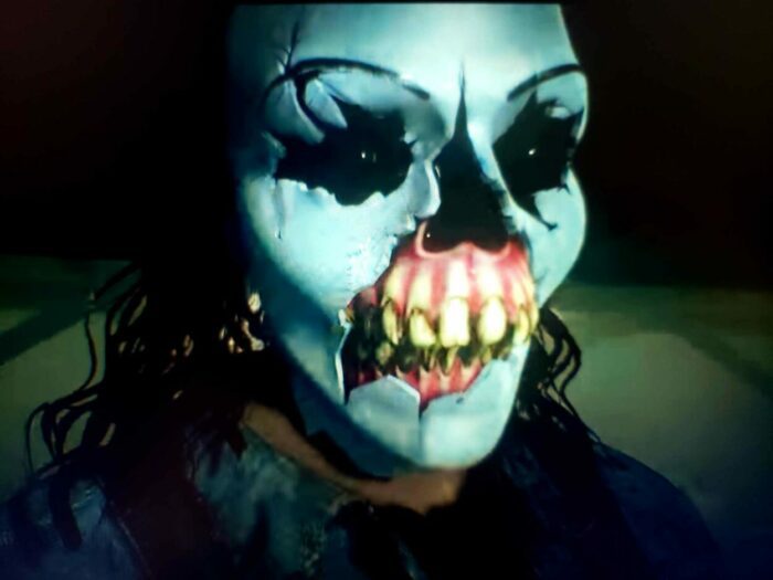 Rami Malek as Joshua Washington in the video game Until Dawn (2015). Screen capture. ©2015 Sony Computer Entertainment. A long haired man in a terrifying all white mask featuring a mouth with no lips stands staring with his eyes aglow.