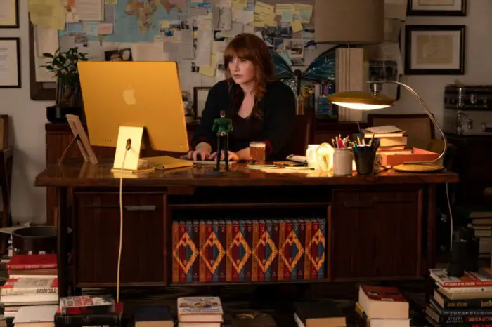 Bryce Dallas Howard as Elly Conway in Argylle, directed by Matthew Vaughn. Universal Pictures; Apple Original Films; and MARV. Red haired author Elly Conway sits at a desk writing, the wall behind her covered in various sticky notes, maps, and novel details.
