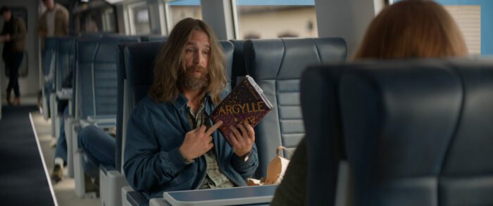 Sam Rockwell as Aidan in Argylle, directed by Matthew Vaughn. Universal Pictures; Apple Original Films; and MARV. Bearded, long haired Aidan sits on a train reading a copy of the book Argylle.