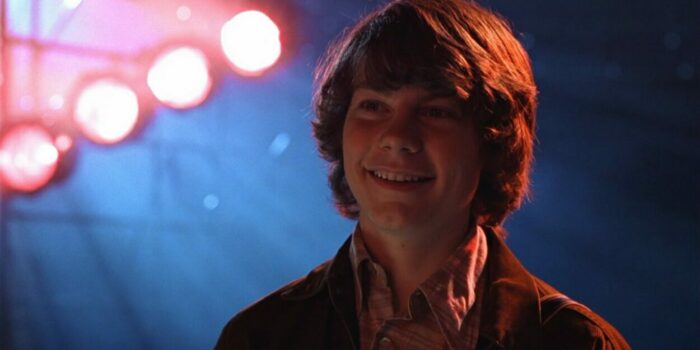 William smiles as he watches Stillwater perform for the first time. Lights are glowing behind him. 