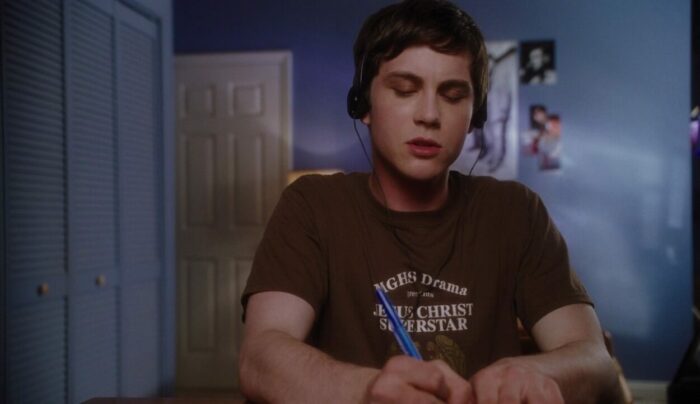 Charlie is sitting at his desk in his room with headphones on, listening to music and writing. 