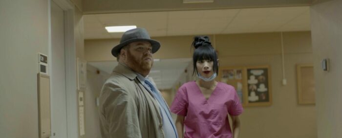 Rod Emmer and Bai Ling as Detective Rod Emmer and Nurse Nancy in The Omicron Killer (2024). © Dark Knites Entertainment. Nurse in pink scrubs and a detective in a long trench coat and fedora idly chatting in a hospital hallway.