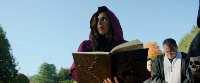 Felissa Rose and Daved Street as the Necromancer and Father Death in The Omicron Killer (2024). © Dark Knites Entertainment. The Necromancer wearing a hooded red robe and holding a strange book reads in the cemetery, while her acolytes stand nearby.
