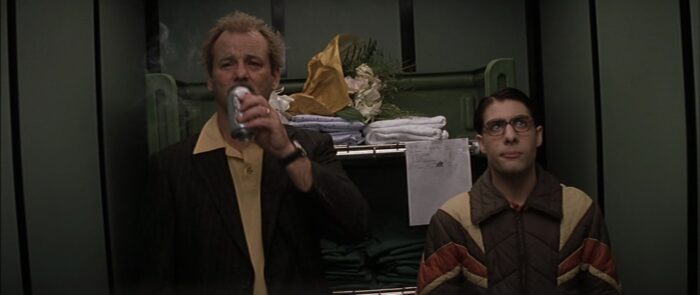 A disheveled Herman Blume (Bill Murray) smoking a cigarette and drinking a diet coke in an elevator with Max Fischer (Jason Schwartzman) in his signature glasses and a jacket.