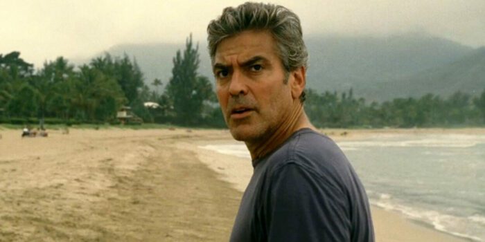 Standing on  a beach, George Clooney) turns and faces the camera in The Descendants.