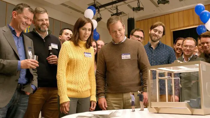 As their colleagues watch and smile, Audrey (Kirsten Wiig) and Paul Sefranek (Matt Damon) stare at "downsized" tiny people waving to them in Downsizing.
