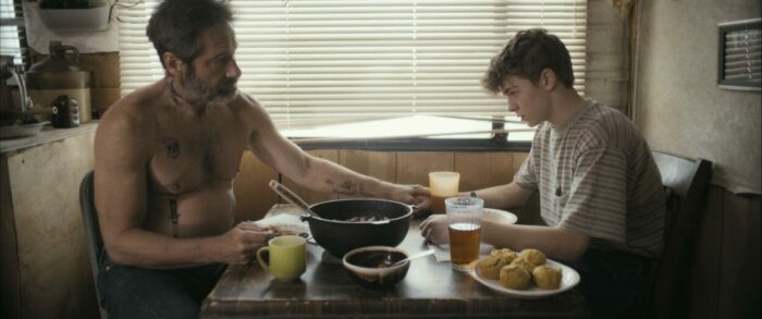 A shirless man at a kitchen table reaches to put his hand on a teen's hand in Adam the First.