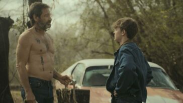 A shirtless man with tattoos talks to a teen in a denim jacket next to a car in Adam the First