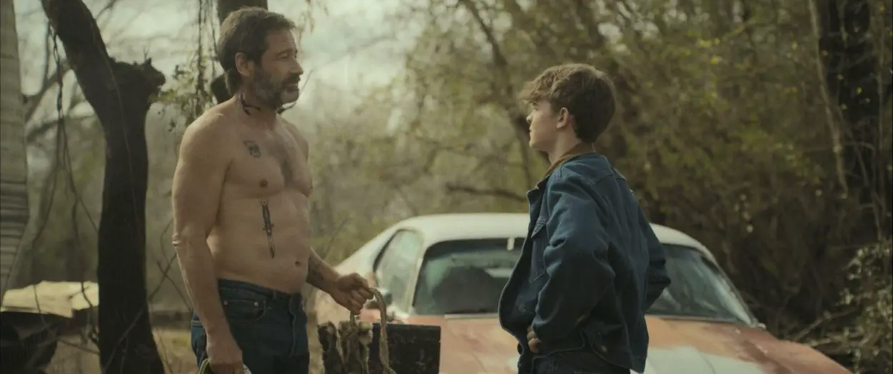 A shirtless man with tattoos talks to a teen in a denim jacket next to a car in Adam the First