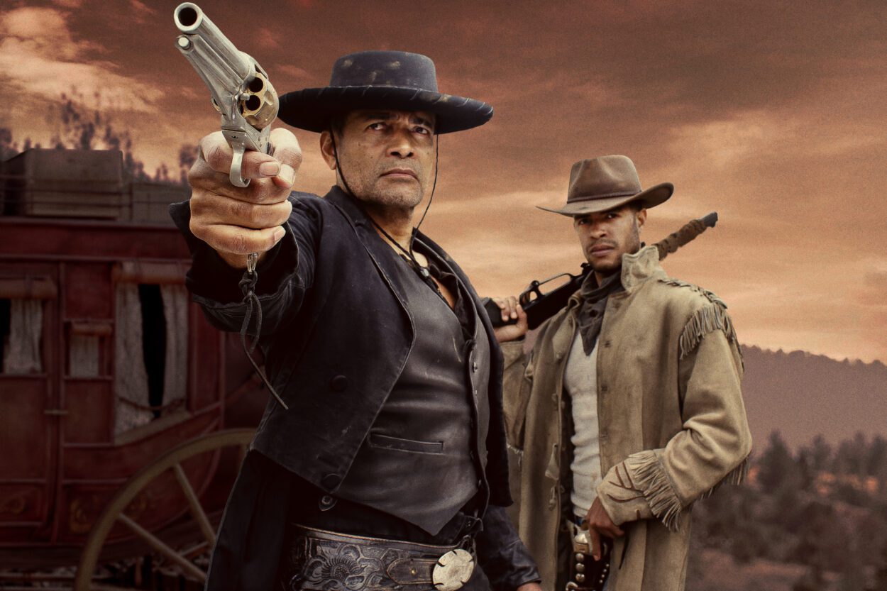 Two men dressed like cowboys stand in front of a stagecoach and an orange sky. The man in front points a gun off screen in Outlaw Posse.