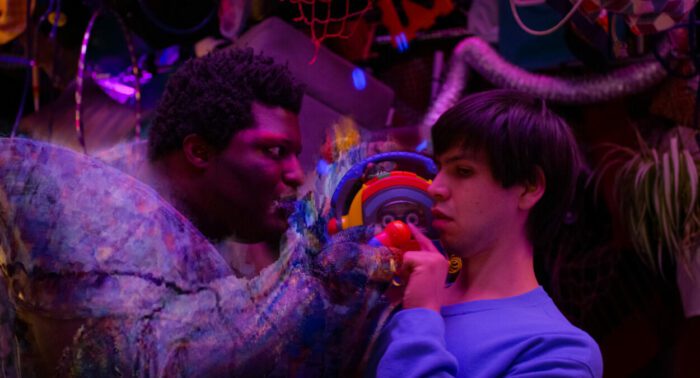 (L-R) Larry Owens, Julio Torres as Craigslist and Alejandro in Problemista (2024) Credit: Courtesy of A24. The living embodiment of Craigslist interacts with the mousy young Alejandro in a surreal moment bringing the internet site to life.