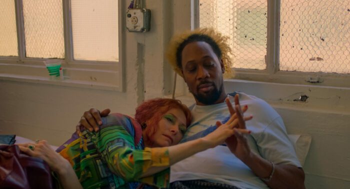 (L-R) Tilda Swinton, RZA as Elizabeth and Bobby in Problemista (2024). Credit: Courtesy of A24. Interracial husband and wife in bed together, clothed and holding hands intimately.