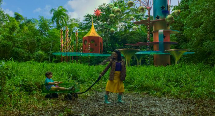 (L-R) Logan J. Alarcon-Poucel and Catalina Saavedra as young Alejandro and Dolores in Problemista (2024). Credit: Courtesy of A24. A mother pulls her small son in a wagon through a magical forest, a wondrous playground in the background behind them.