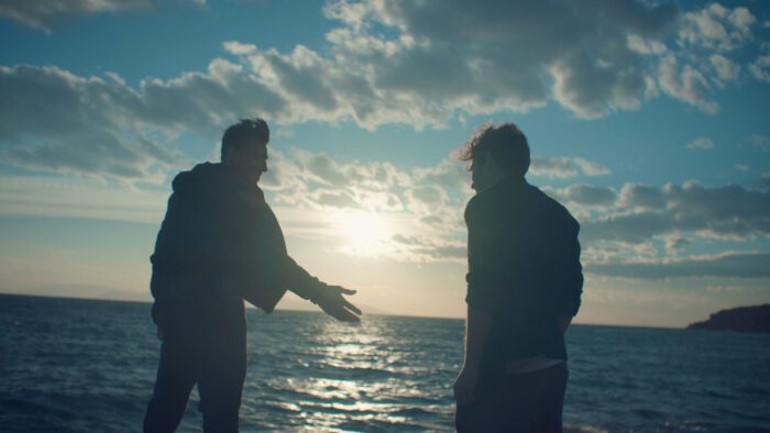 Aenas and Alex are depicted in near-silhouette in front of the ocean sunset.