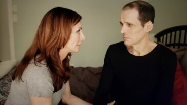 Ena O'Rourke and Joey Collins as Carol and Jack in There is a Monster (2024). Screen capture off of Vimeo access courtesy of Gravitas Features. A red-haired woman sits on the bed consoling a frightened man in a black shirt.