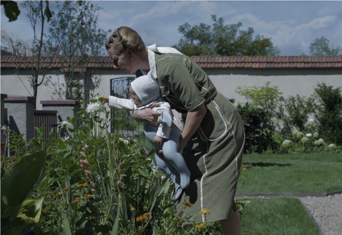 Sandra Hüller, playing Hedwig Höss in Glazer's The Zone of Interest, holds a child up to a flower in a garden. 