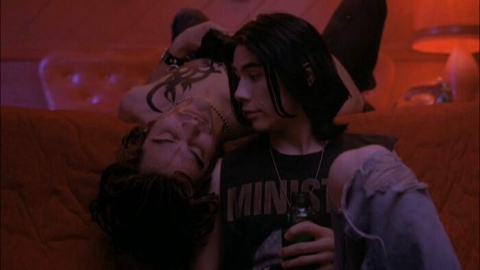 Jordan White (James Duval) and Xavier "X" Red (Jonathon Schaech) in The Doom Generation. One is lying with their head hanging off the edge of the bed. The other is seated on the floor next to him, face turned close to his.