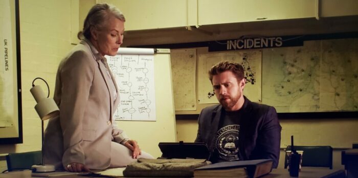 Rowena Bentley and Derek Nelson as Julia Goldstein and Jim Peters in Gods of the Deep (2024). Screen capture off of Vimeo. An older woman and a young man sit at a desk watching a tablet displaying strange images of ruins on the ocean floor.