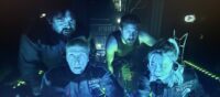 [L to R] Rory Wilton, Tim Cartwright, Derek Nelson, Makenna Guyler as Hank, Gordon, Jim, and Christine in Gods of the Deep (2024). Screen capture off of Vimeo access courtesy of Quiver Distribution. Crew of an experimental submersible gathered around the main controls, their faces lit blue by glowing electronics, all looking worried.