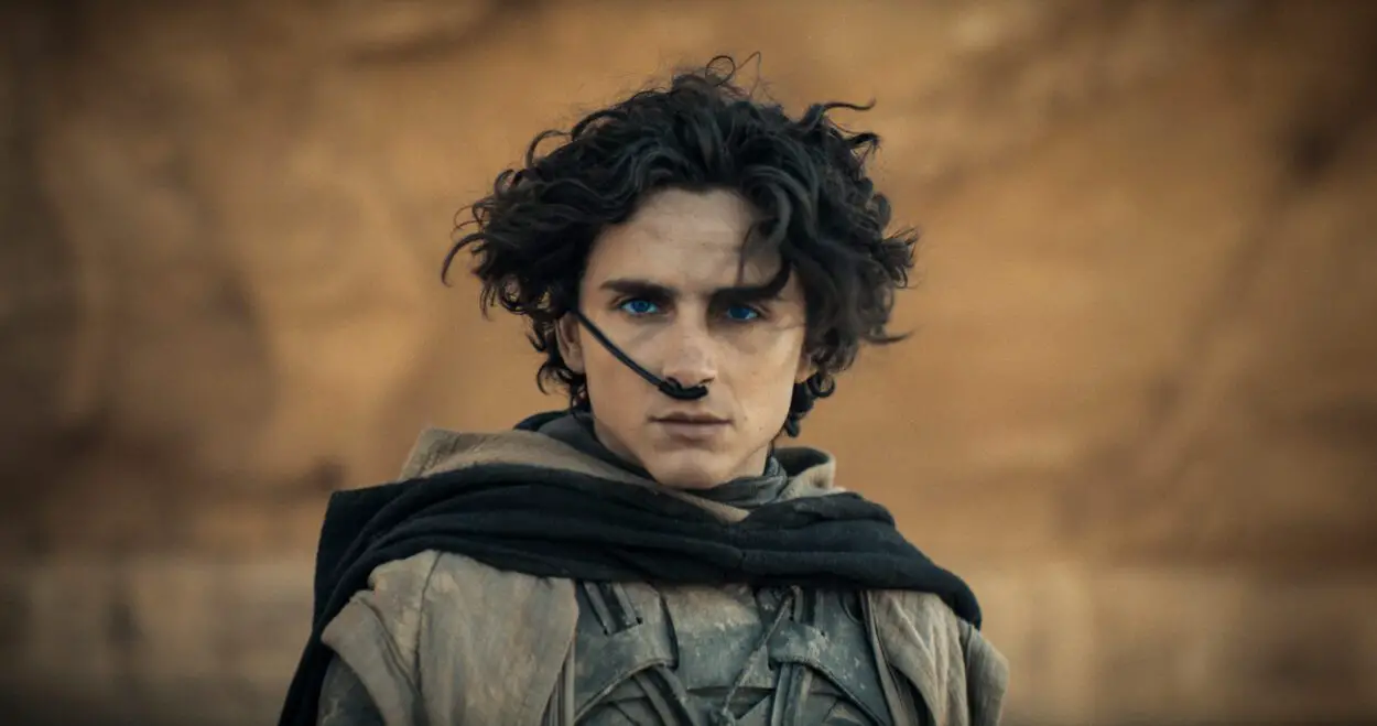 TIMOTHÉE CHALAMET as Paul Atreides in Warner Bros. Pictures and Legendary Pictures’ action adventure “DUNE: PART TWO,” a Warner Bros. Pictures release. © 2023 Warner Bros. Entertainment Inc. All Rights Reserved. Photo Credit: Courtesy Warner Bros. Pictures. Paul standing before a rock wall, breathing through a nose tube, wearing the desert gear of the Fremen.