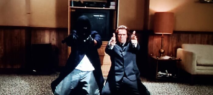 David Della Rocco and Willem Dafoe as Rocco and Agent Smecker in The Boondock Saints (1999). Screen capture off of DVD. Franchise Pictures. Rocco in long coat and ski mask on his knees firing pistols with Agent Smecker mirroring his actions as a flashback plays out; Smecker essentially living in his conclusions about a crime scene.