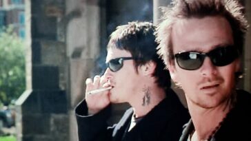 Norman Reedus and Sean Patrick Flannery as Murphy and Connor MacManus in The Boondock Saints (1999). Screen capture off DVD. Franchise Pictures. Two brothers in matching jeans and black shirts wearing sunglasses while smoking.