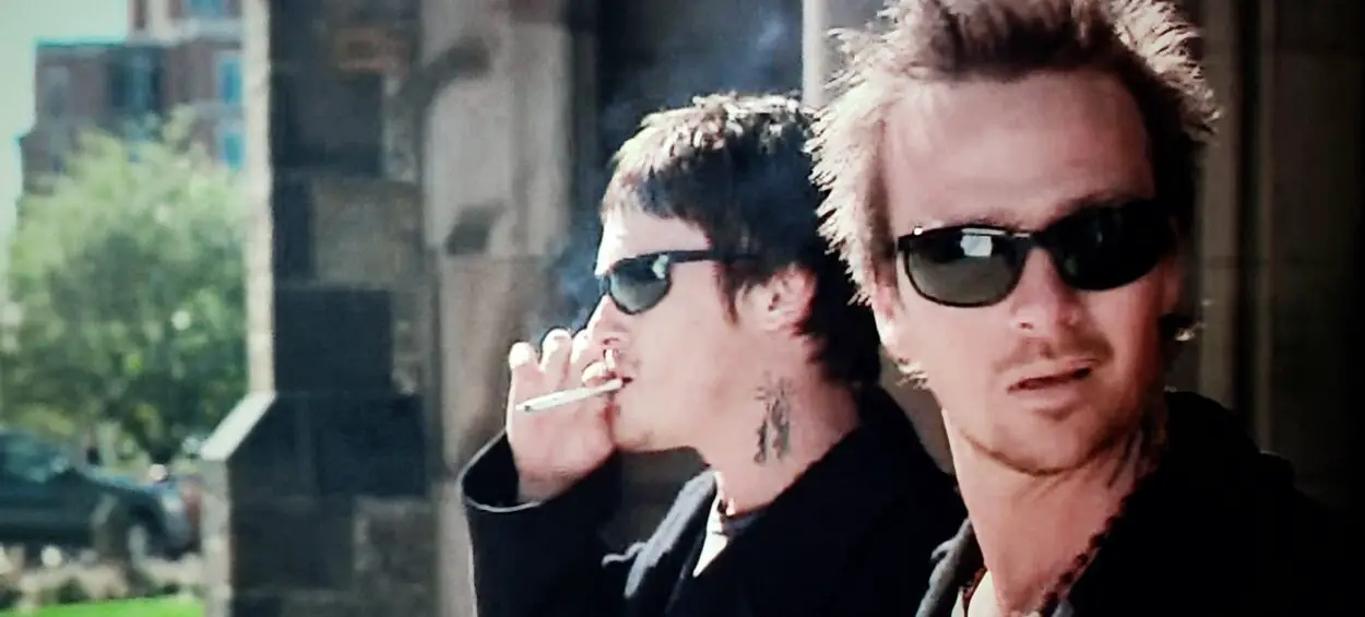 Norman Reedus and Sean Patrick Flannery as Murphy and Connor MacManus in The Boondock Saints (1999). Screen capture off DVD. Franchise Pictures. Two brothers in matching jeans and black shirts wearing sunglasses while smoking.
