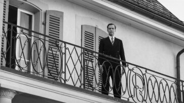 Michael Neuenschwander as Heinreich Zwygart in A Forgotten Man (2022). Sovereign Films. Black and white scene of a Swiss man standing alone on a balcony looking down.