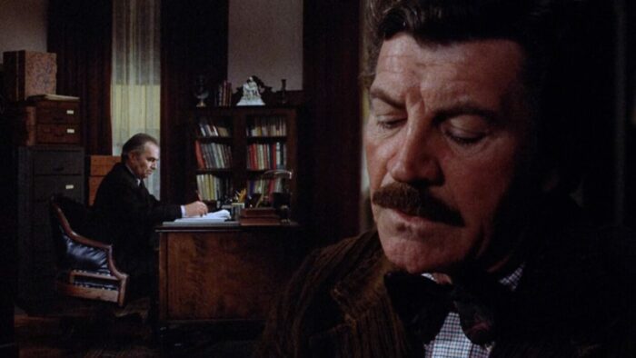 Two men in a split-diopter shot. The man in the background sits at a desk and the man in the foreground is close on his face.