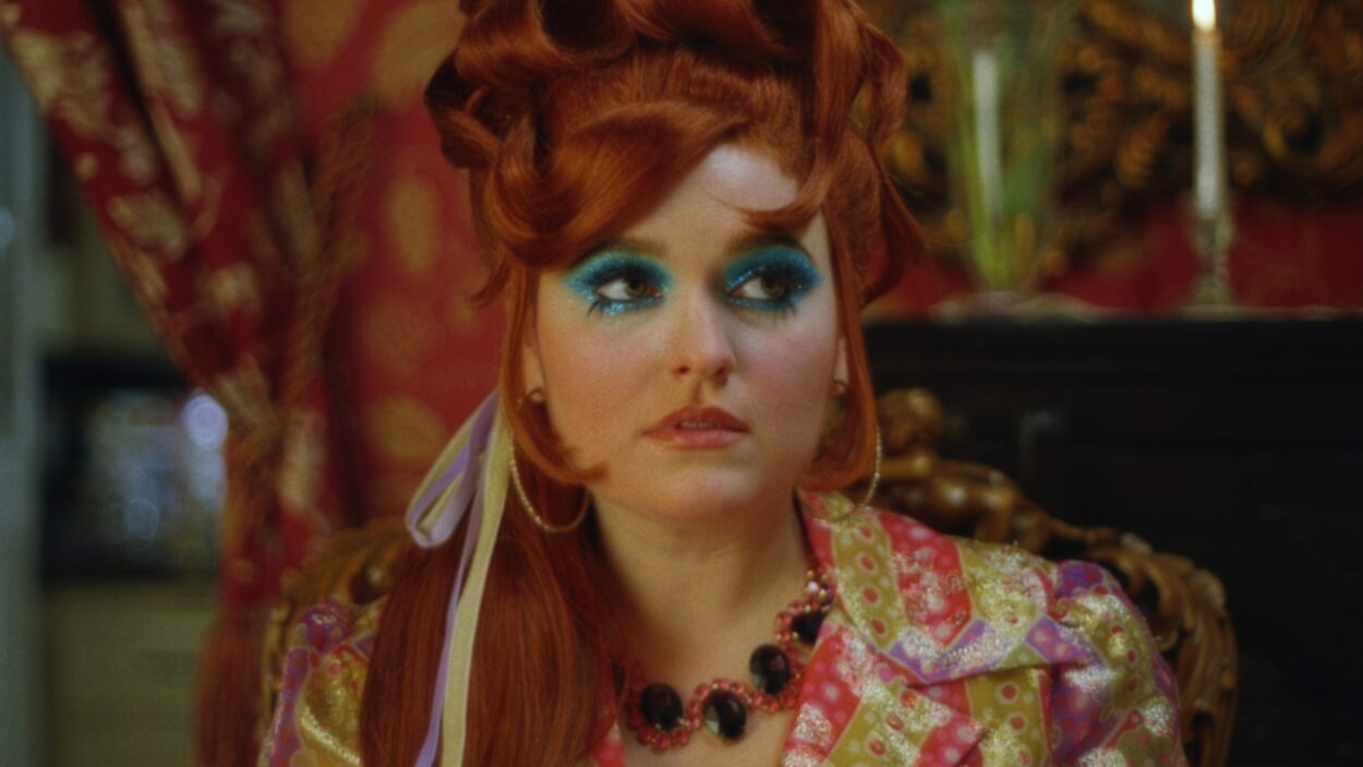 Scarlet Moreno as Velma in the short film Velma (2022). Velma standing alone dressed in a campy 1960s style with her hair done up in lavish orange curls.