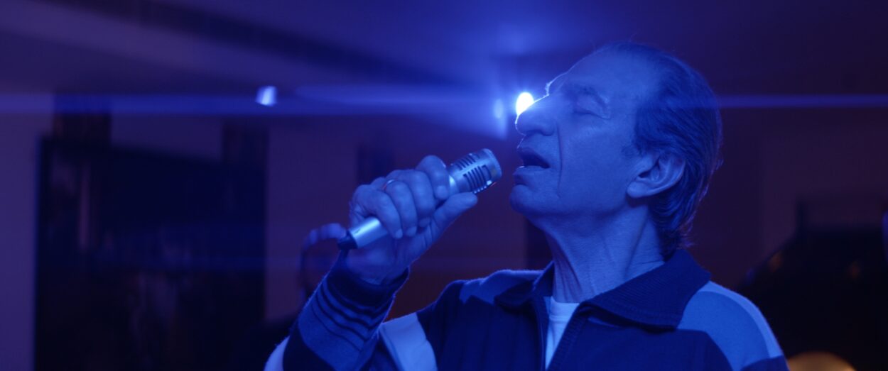 Sasson Gabay as Meir sings into a microphone in Karakoe, he and his surroundings bathed in purpole light.