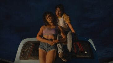 Jackie, wearing a purple tank top, and Lou, wearing a flannel and white t-shirt, sit and lean atop a car at night in Love Lies Bleeding