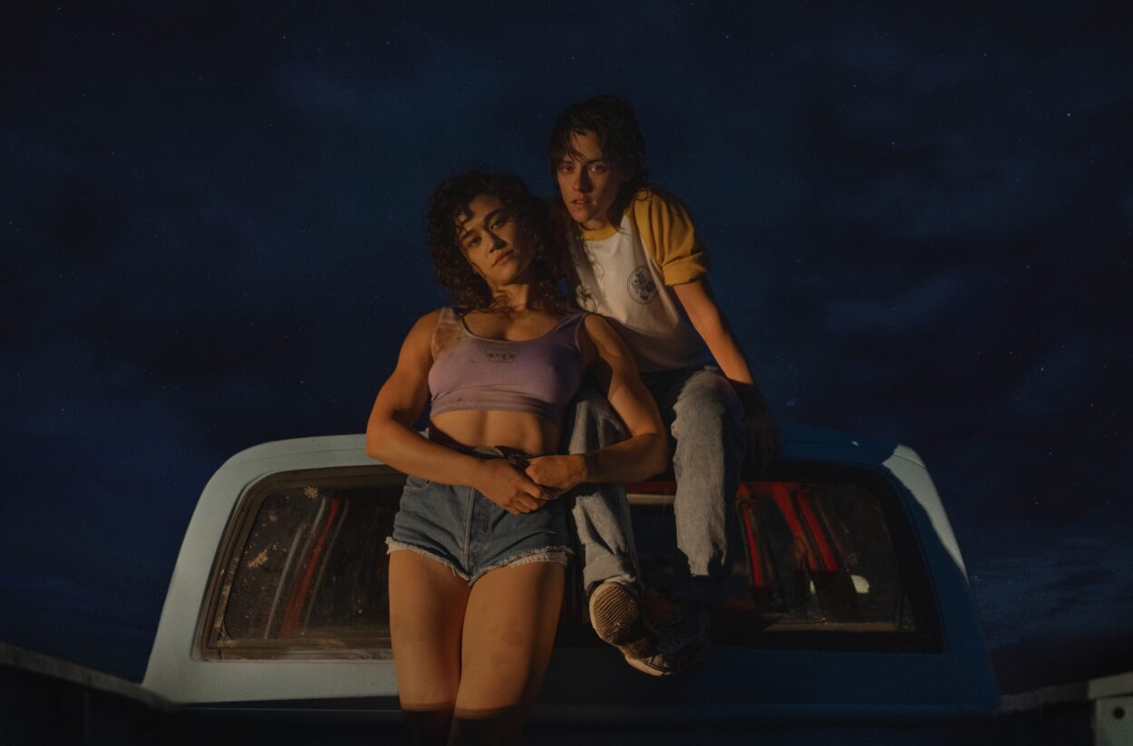 Jackie, wearing a purple tank top, and Lou, wearing a flannel and white t-shirt, sit and lean atop a car at night in Love Lies Bleeding