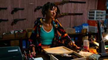 Patricia Allison as Ella Patchet in Paradise (2023). Young black woman at work behind the counter in a gun store, a wall of firearms behind.