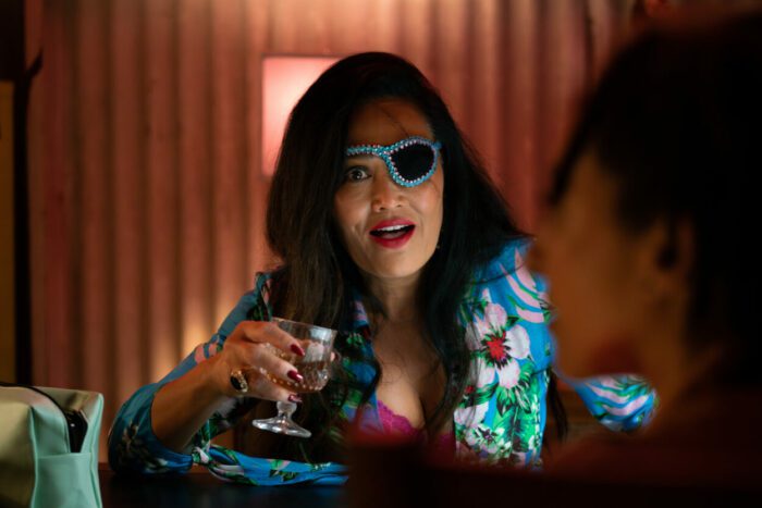 Tia Carrere as Lee Paige in Paradise (2023). Underworld crime boss Lee Paige sits at her desk sipping brandy while wearing a colorful eyepatch and floral dress.