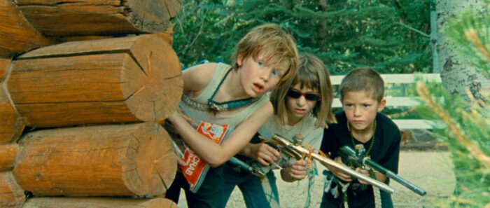 Three kids look around the corner of a log cabin armed with a paintball gun in Riddle of Fire
