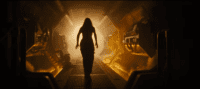 A woman, her stance fearful, creeps down a the corridor of a space station, fire lighting the path ahead of her, she is surrounded in darkness
