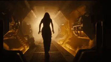 A woman, her stance fearful, creeps down a the corridor of a space station, fire lighting the path ahead of her, she is surrounded in darkness
