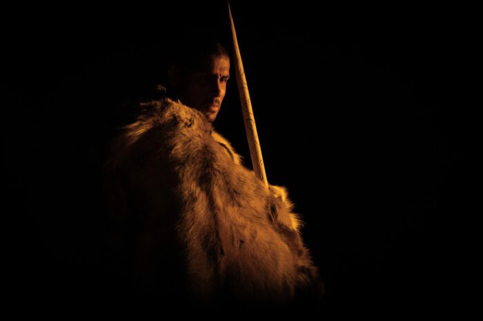Chuku Modu as Adem in Bleecker Street's OUT OF DARKNESS. Credit: Bleecker Street. A stone age man lit by a campfire stands in the dark wearing furs and carrying a spear.