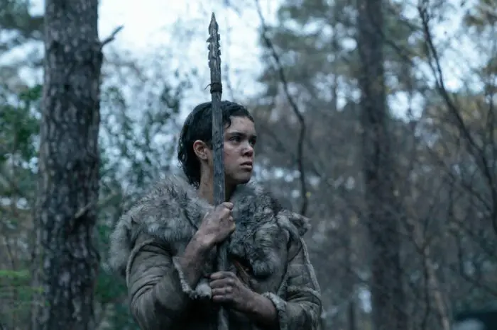 A young woman in stone age clothes composed of furs stands alone in a grim forest holding a spear in Out of Darkness